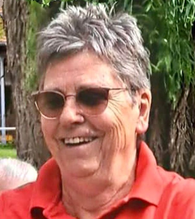 Eine lächelnde Person mit Sonnenbrille und rotem Hemd steht im Freien in der Nähe eines Baumes.