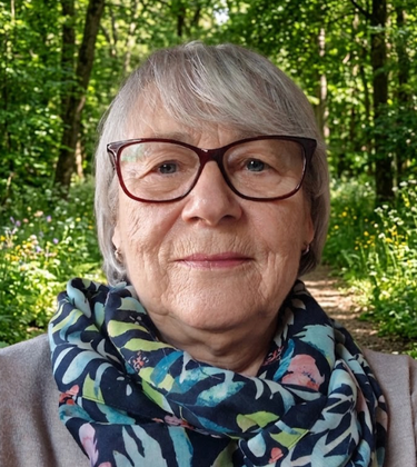 Porträt einer Person mit Brille und blauem Blumenschal auf einem sonnenbeschienenen Waldweg