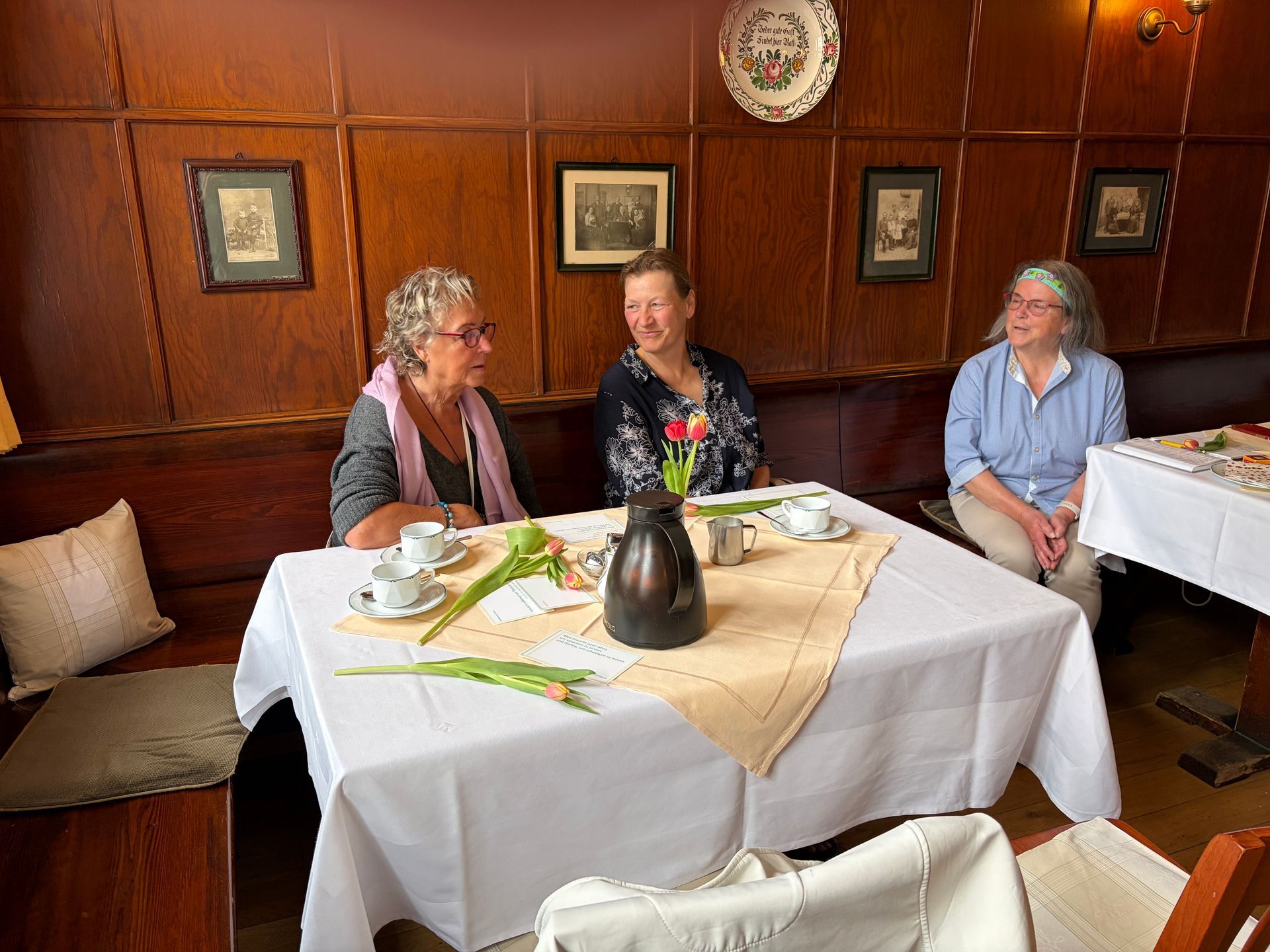 Drei Frauen sitzen an einem Restauranttisch mit Getränken und einer weißen Tischdecke in einem holzgetäfelten Speisesaal.