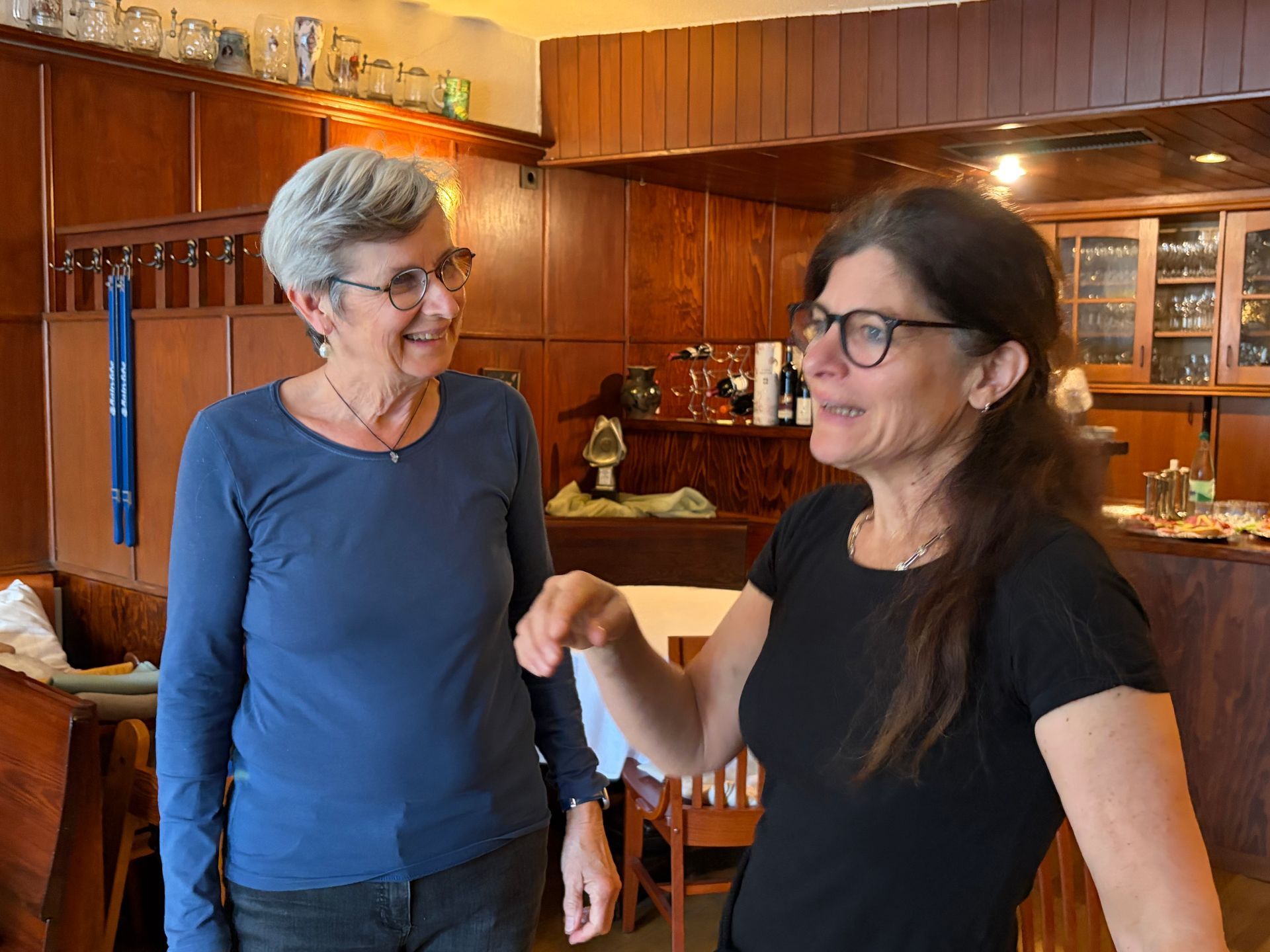 Zwei Frauen unterhalten sich in einem holzgetäfelten Zimmer, eine gestikuliert dabei, während sie lächeln.