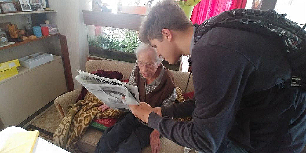 Das Bild zeigt eine berührende Alltagsszene: Ein junger Mann beugt sich aufmerksam zu einer älteren Dame herab, die auf einem Sofa sitzt. Gemeinsam halten sie eine weit geöffnete Zeitung – den ‚Odenwälder‘ – und lesen darin. Die Seniorin trägt eine Brille und blickt konzentriert auf die Nachrichten, während der junge Helfer ihr mit einer ruhigen, zugewandten Körperhaltung beisteht. Im Hintergrund ist eine gemütliche Wohnzimmeratmosphäre mit Pflanzen und einem Aquarium zu erkennen. Dieses Motiv verkörpert perfekt unsere Mission: Wir schlagen die Brücke zwischen den Generationen und sorgen dafür, dass unsere Mitglieder informiert und am gesellschaftlichen Leben beteiligt bleiben.
