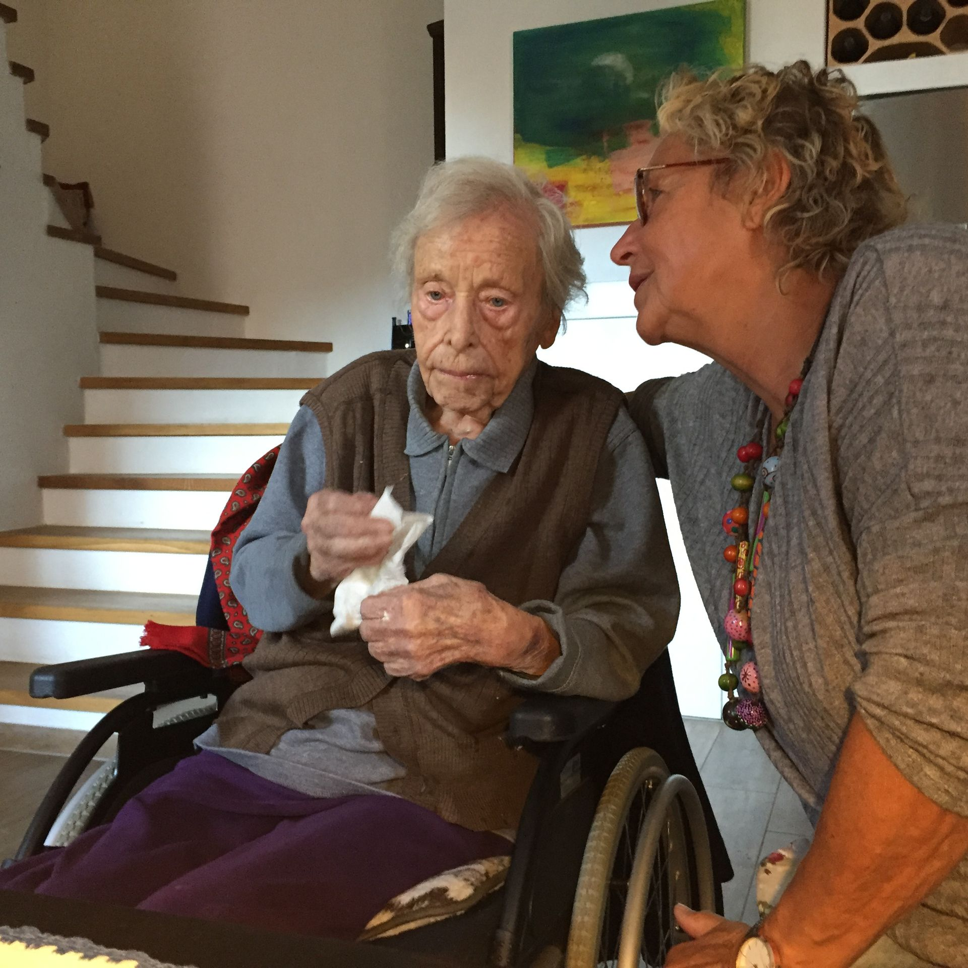 Dieses Foto fängt einen sehr privaten und intensiven Moment der Seniorenhilfe ein. Es zeigt eine Helferin, die sich einer älteren, sichtlich geschwächten Dame liebevoll zuneigt. Die Helferin, eine Frau mit Brille und Locken, blickt ihr Gegenüber mit einem Ausdruck von tiefer Empathie und Ruhe an. Die Seniorin wirkt zerbrechlich, doch die Nähe der Helferin vermittelt Sicherheit und Trost. Im Hintergrund ist eine helle Treppe und ein farbenfrohes Bild zu sehen, was die häusliche, private Atmosphäre unterstreicht. Dieses Bild steht für das, was kein Formular ausdrücken kann: Wir schenken Zeit, wir hören zu und wir lassen niemanden in seiner Einsamkeit allein.