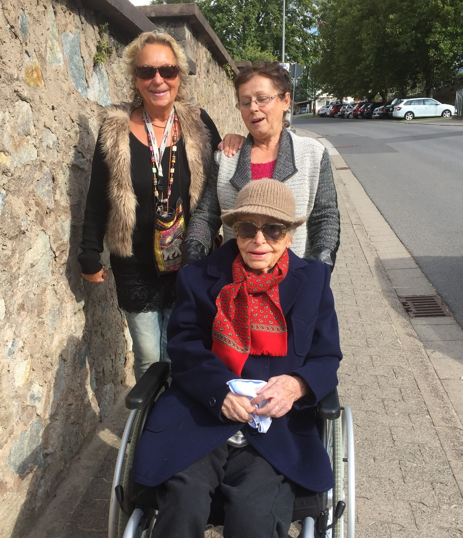 „Das Bild zeigt drei Damen bei einem gemeinsamen Spaziergang an einem sonnigen Tag in Groß-Umstadt. Links im Bild lächelt eine Frau mit blonder Mähne und Sonnenbrille in die Kamera; sie trägt eine markante Weste in Fell-Optik und eine bunte Ethno-Tasche. In der Mitte steht eine weitere Helferin, die ihre Hand freundlich auf die Schulter der Seniorin legt, die im Vordergrund sitzt. Die Seniorin trägt einen schicken Hut, eine Sonnenbrille und einen roten Schal. Sie befinden sich auf einem Gehweg neben einer rustikalen Natursteinmauer, im Hintergrund säumen grüne Bäume und geparkte Autos die ruhige Straße. Die Szene strahlt Lebensfreude, Zusammenhalt und die Freude am gemeinsamen Unterwegssein aus.