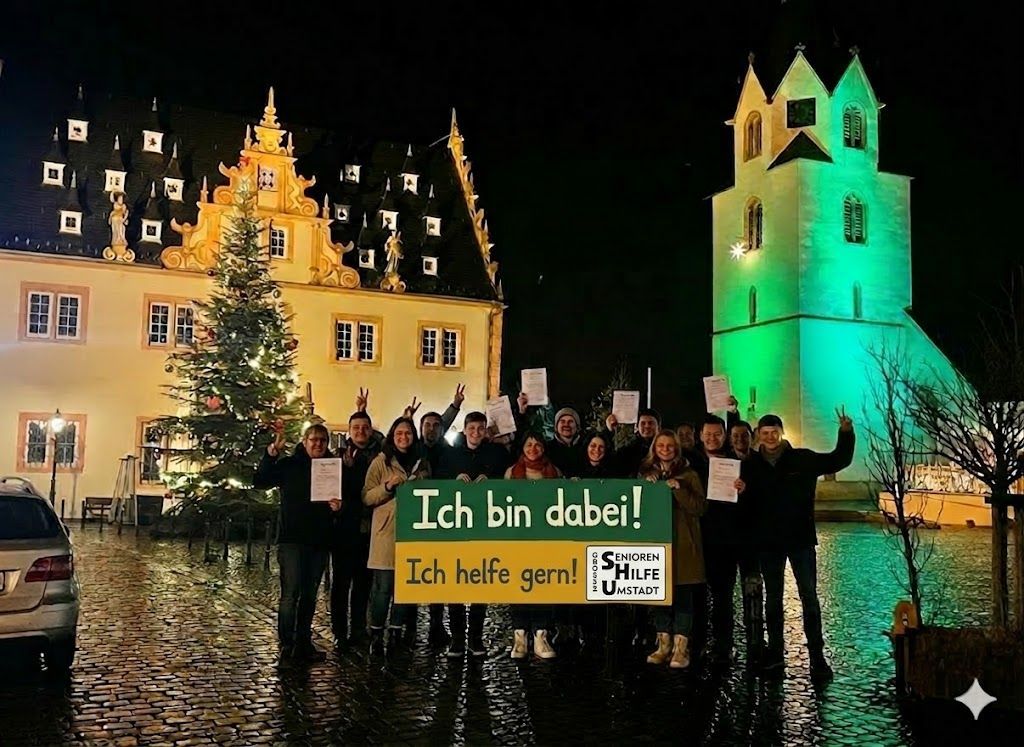 Das Bild zeigt eine große, fröhliche Gruppe von Menschen, die sich nachts auf dem Marktplatz von Groß-Umstadt versammelt hat. Im Hintergrund erstrahlt das prächtige, historisch beleuchtete Rathaus und die Stadtkirche, die in ein festliches grünes Licht getaucht ist. Ein geschmückter Weihnachtsbaum bringt zusätzliche Wärme in die Szene. Die Gruppe hält stolz ein großes Banner in den Händen, auf dem in kräftigem Blau und Gelb steht: ‚Ich bin dabei! Ich helfe gern!‘ Viele halten zudem ihre Beitrittserklärungen in die Kamera und zeigen das Friedenszeichen. Dieses Bild sprüht vor Lebensfreude und zeigt deutlich: Wir sind viele, wir sind motiviert und wir sind stolz auf unsere Stadt und unseren Verein