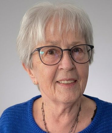 Porträt einer älteren Frau mit kurzem, weißem Haar, Brille und blauem Pullover, die vor einem schlichten Hintergrund sanft lächelt.