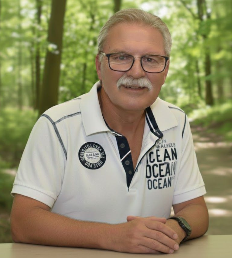 Das Bild zeigt das Porträt von Friedrich Koch, einem Mann mit einer herzlichen und vertrauenswürdigen Ausstrahlung. Er hat kurzes, helles Haar und trägt einen gepflegten grauen Schnurrbart. Mit seiner modernen Brille und dem sportlichen, weiß-blau gestreiften Poloshirt wirkt er aktiv und nahbar. Der Hintergrund zeigt eine sanft verschwommene, grüne Waldlandschaft, die Ruhe und Bodenständigkeit vermittelt. Es ist das Bild eines Mannes, der mit beiden Beinen im Leben steht und sich mit voller Kraft für die Senioren in Groß-Umstadt einsetzt.