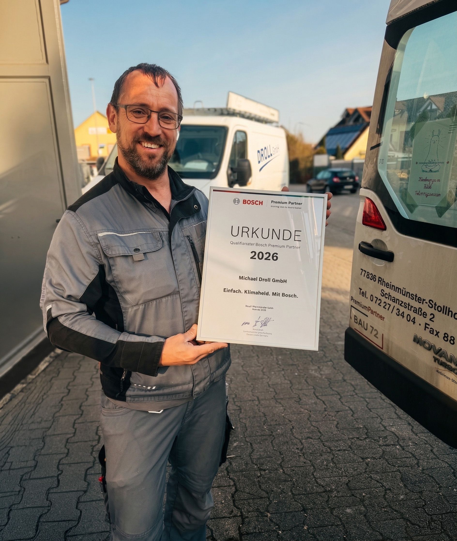 Eine Person steht lächelnd zwischen zwei Lieferwagen auf einem gepflasterten Freigelände und hält eine Urkunde aus dem Jahr 2020 in der Hand.