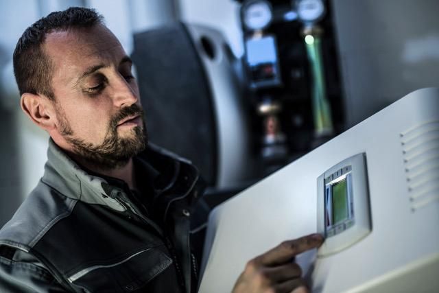 Ein Arbeiter in grauer Arbeitskleidung justiert die Einstellungen an einem an der Wand montierten digitalen Bedienfeld für Industrieanlagen.