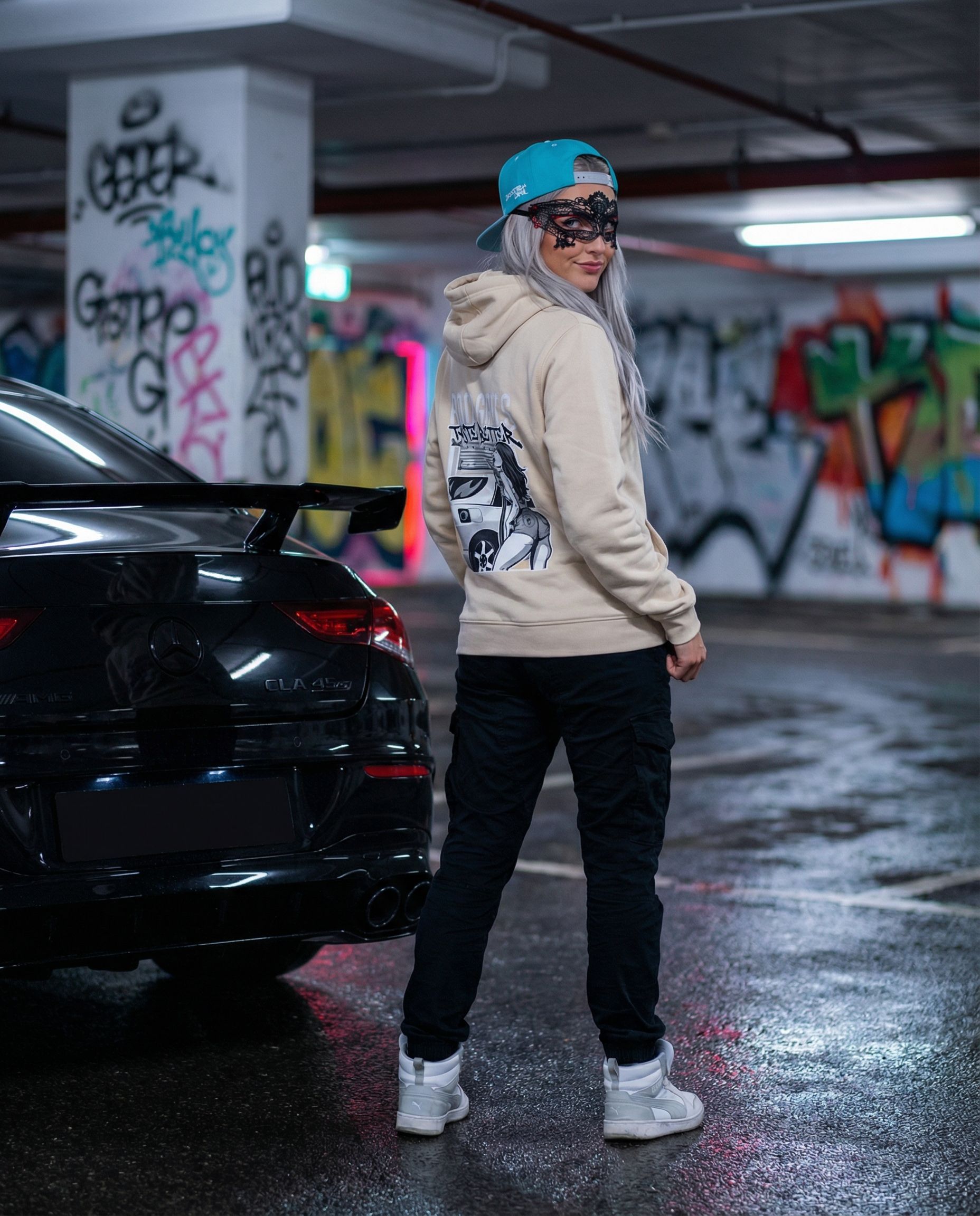 Eine Frau mit Kapuzenpulli und Kappe steht in einem mit Graffiti besprühten Parkhaus neben einem schwarzen Auto.