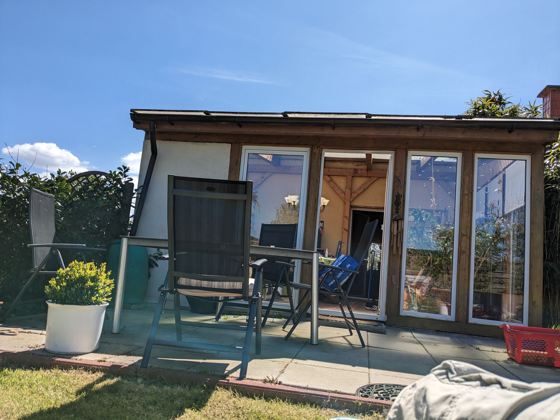 Ein Wintergarten mit Glastüren, einer Terrasse und Gartenmöbeln an einem sonnigen Tag.