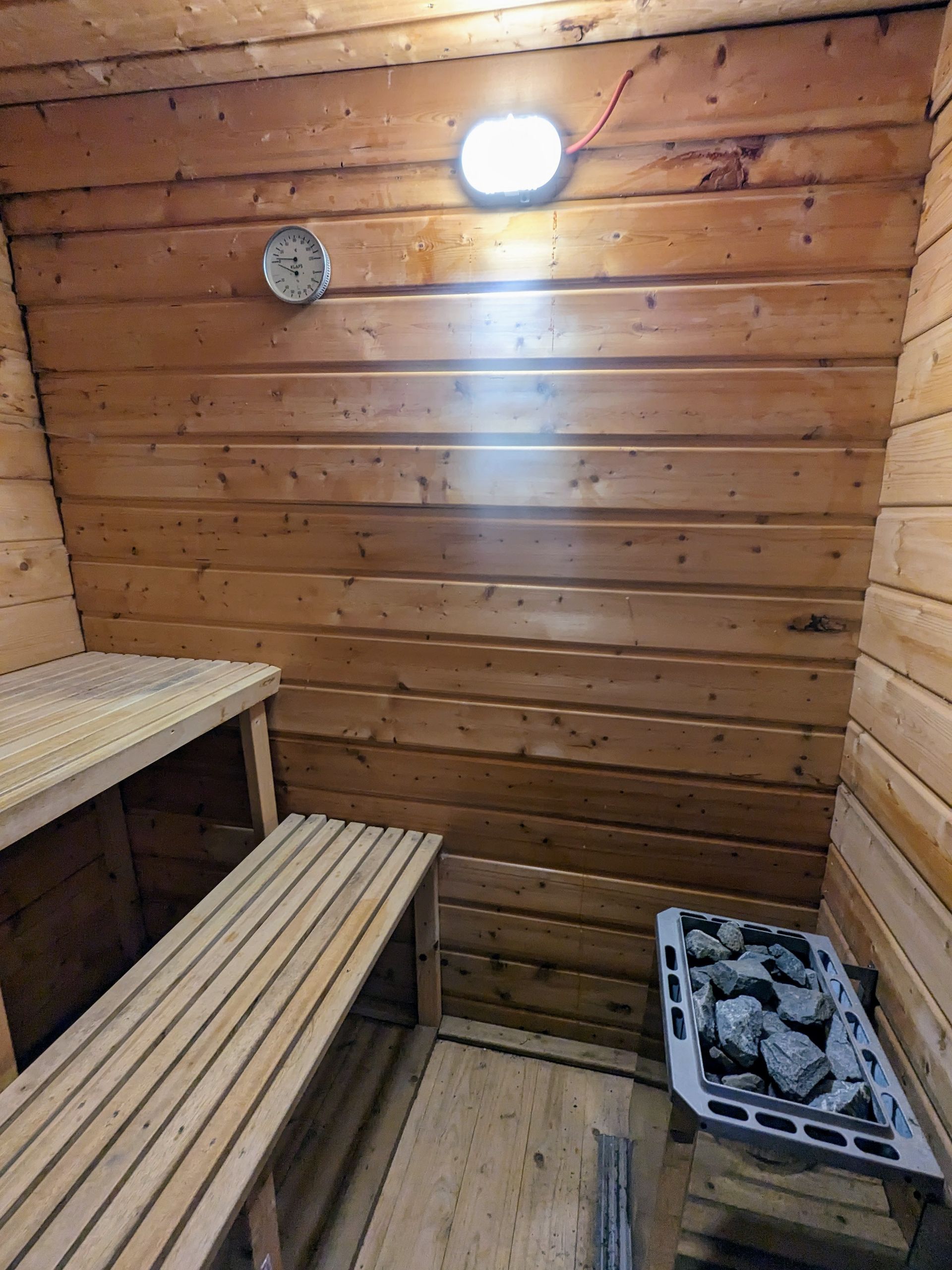 Innenraum einer Holzsauna mit Bänken, Ofen mit Steinen und Deckenlicht.