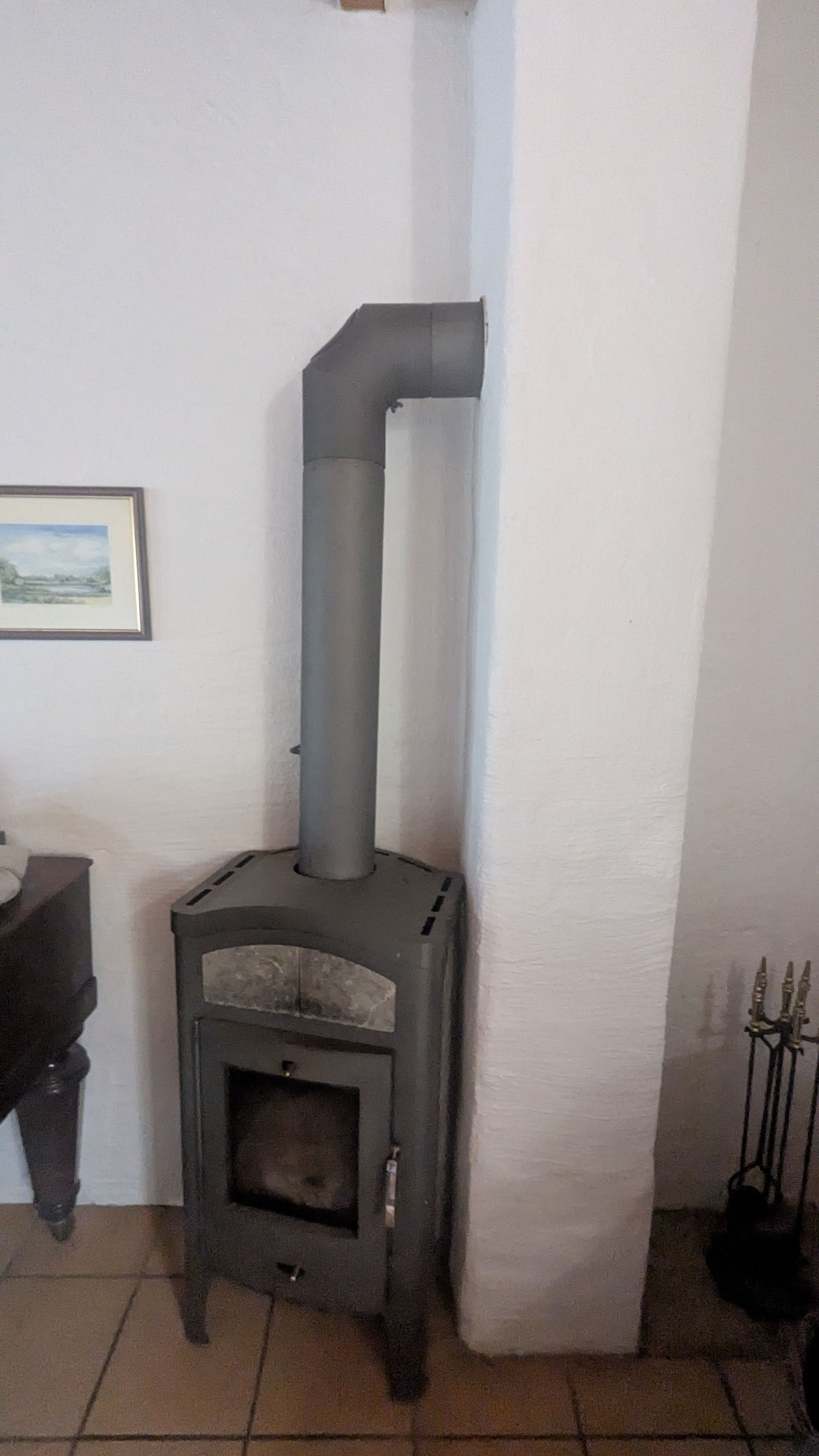 Ein dunkler Holzofen aus Metall mit angeschlossenem Kaminrohr vor einer weißen Wand in einem Raum.