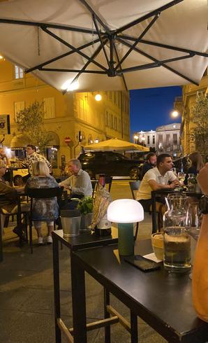 Straßencafé in der Abenddämmerung mit Gästen unter Sonnenschirmen, warmem Licht und Getränken auf Tischen mit Blick auf eine beleuchtete Straße.