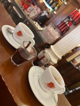Zwei Tassen Kaffee und zwei Gläser Tee auf einem Holztisch in einem Café, im Hintergrund ist die Theke verschwommen zu erkennen.