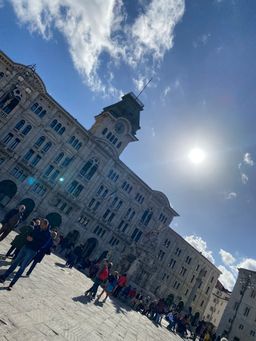 Menschenmenge auf einem sonnenbeschienenen Platz neben einem historischen Uhrturmgebäude unter blauem Himmel