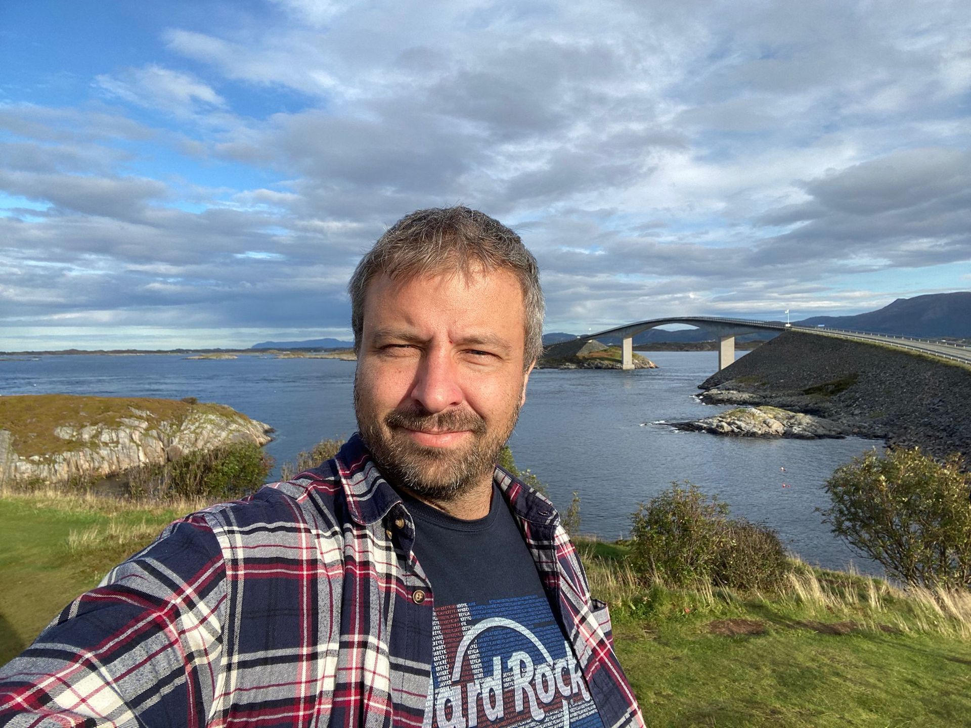 Mann macht ein Selfie auf einer Küstenstraße mit einer Brücke im Hintergrund, blauem Wasser, bewölktem Himmel.
