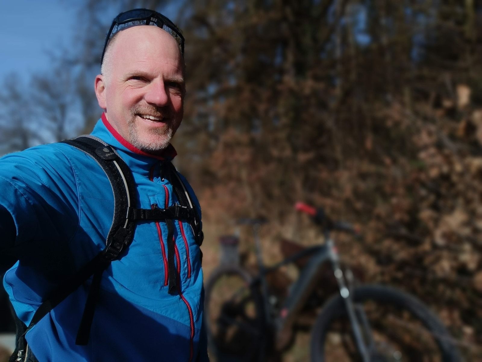 Lächelnder Mann im Freien, mit blauer Jacke, Rucksack und Sonnenbrille, im Hintergrund ein Fahrrad.