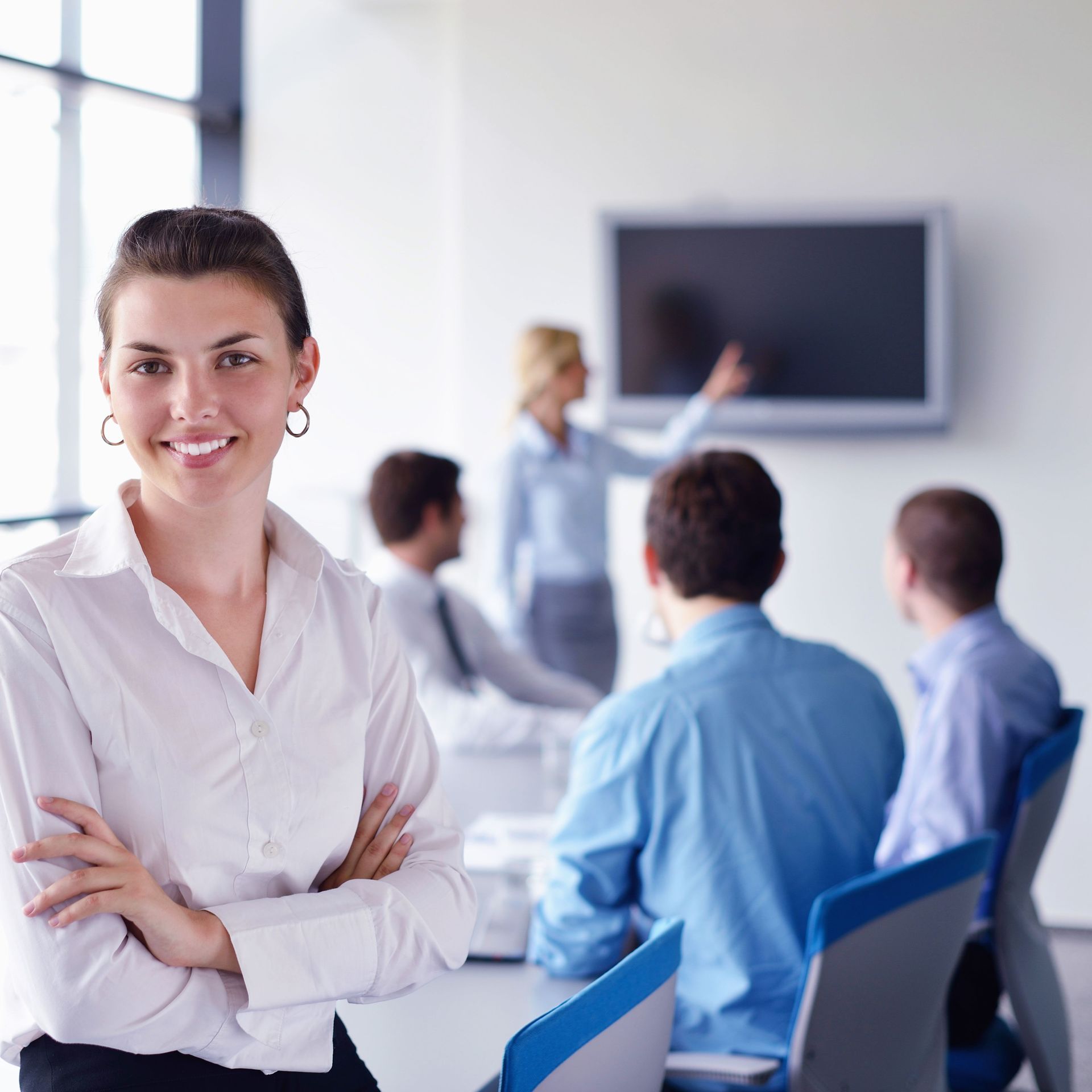 Frau mit verschränkten Armen lächelt in einem modernen Büro. Andere in einer Besprechung, im Hintergrund ein Bildschirm.
