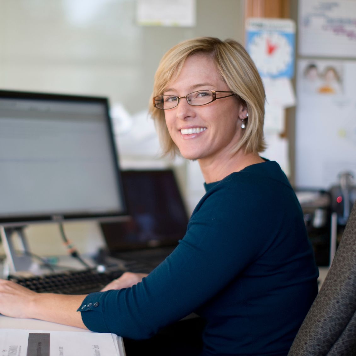 Frau mit blonden Haaren lächelt in die Kamera, während sie an ihrem Schreibtisch arbeitet.