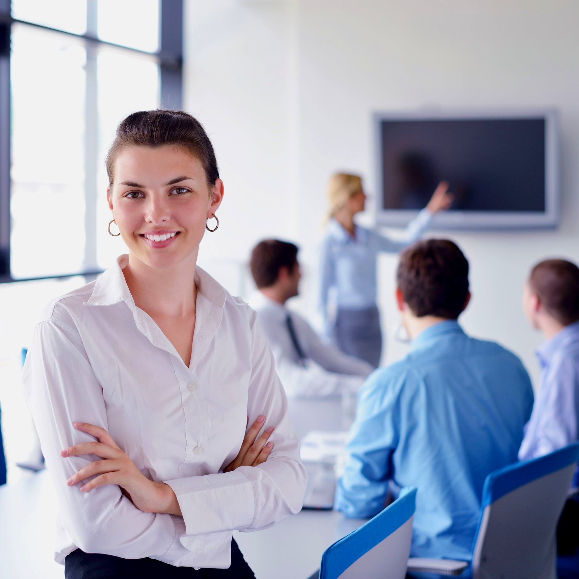Lächelnde Frau mit verschränkten Armen vor einem Konferenzraum. Andere sind im Hintergrund.