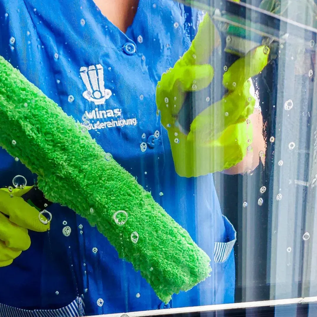 Person in blauer Uniform putzt Fenster mit einem grünen Schrubber und gelben Handschuhen.