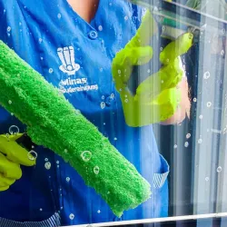 Person in blauer Uniform putzt Fenster mit einem grünen Schrubber und gelben Handschuhen.