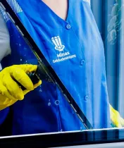 Person mit gelben Handschuhen und blauer Weste reinigt ein Fenster mit einem Abzieher. Auf der Weste ist das Logo „Minas Gebäudereinigung“ zu sehen.