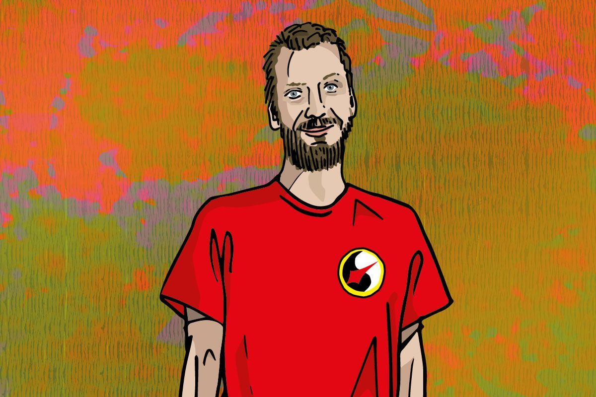 Mann in rotem Hemd mit Logo, lächelt vor einem orangefarbenen, grünen und gelben Hintergrund.