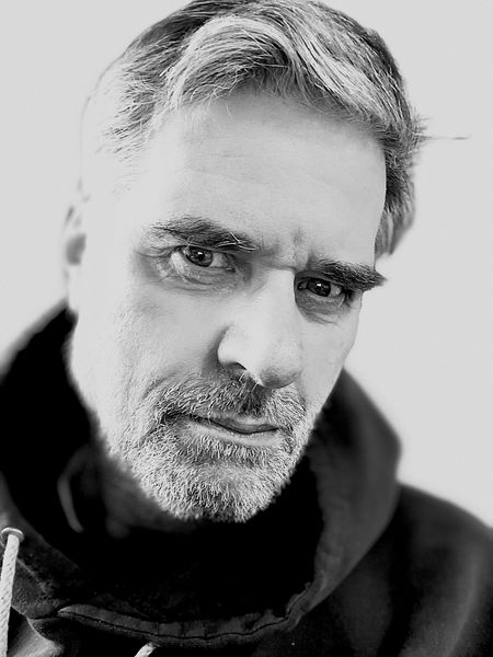 Ein Mann mit ergrauendem Haar und Bart blickt direkt in die Kamera; er trägt einen Kapuzenpullover.