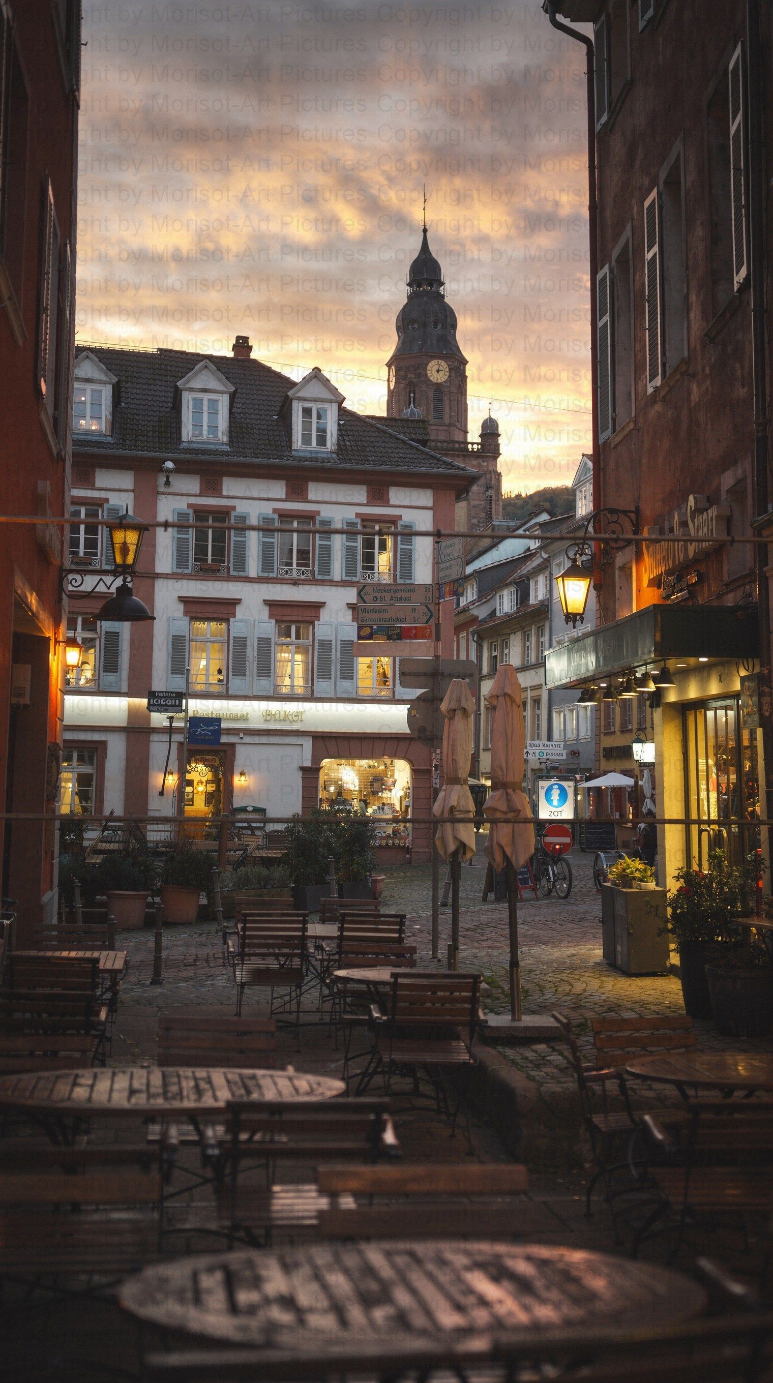 Kopfsteinpflasterstraße in Heidelberg, Deutschland, mit Außengastronomie, Geschäften, Gebäuden und einem Kirchturm. Abendbeleuchtung.