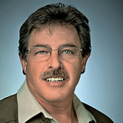 Porträt eines Mannes mit braunen Haaren, Brille und Schnurrbart, der vor blauem Hintergrund lächelt.