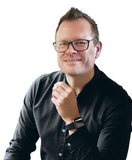 Eine Person mit Brille und schwarzem Hemd lächelt, während sie sich die Hand ans Kinn hält; aufgenommen vor einem weißen Hintergrund.