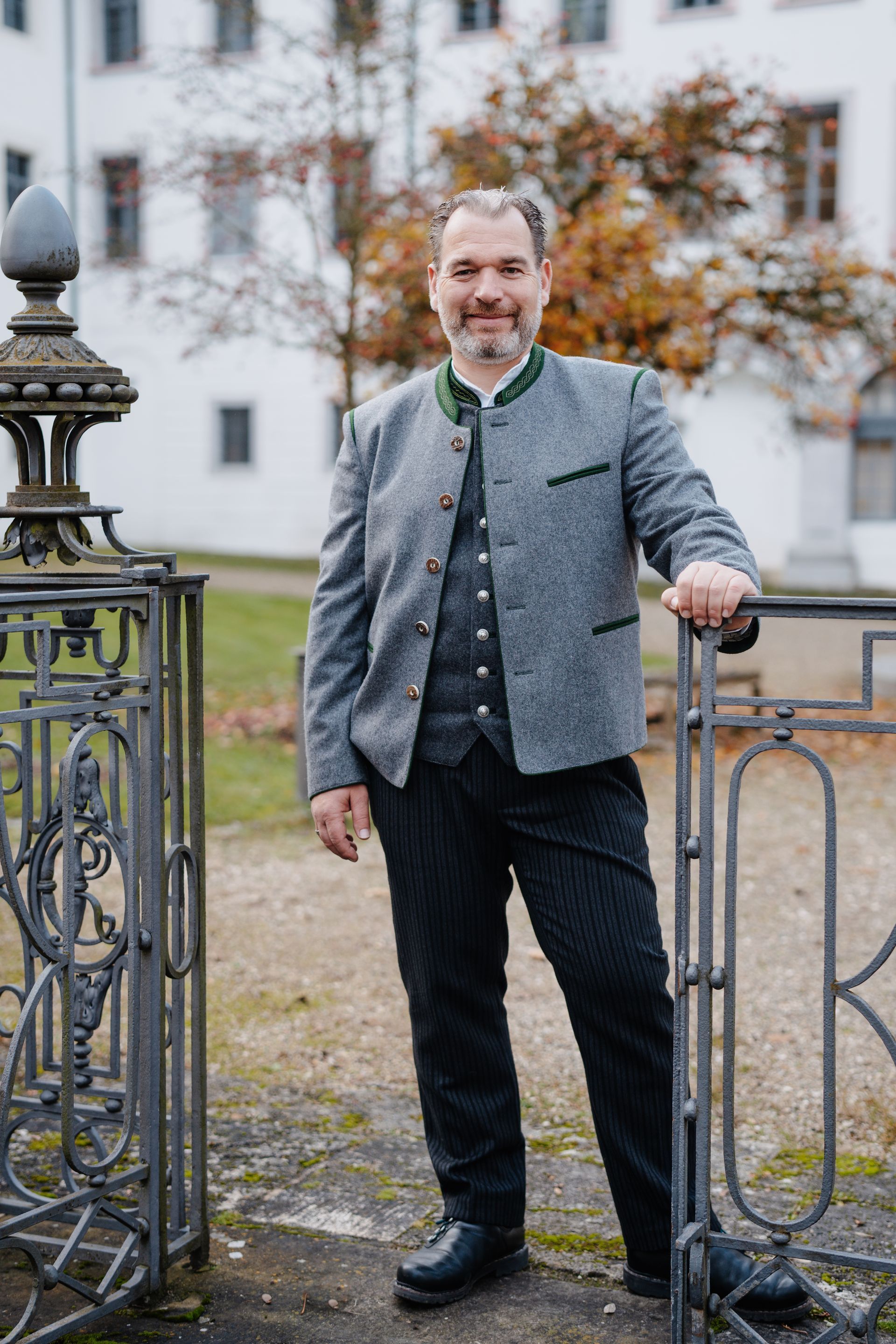 Matthias Plattner in traditioneller Kleidung lehnt an einem schmiedeeisernen Tor, im Hintergrund ist ein Gebäude zu sehen.