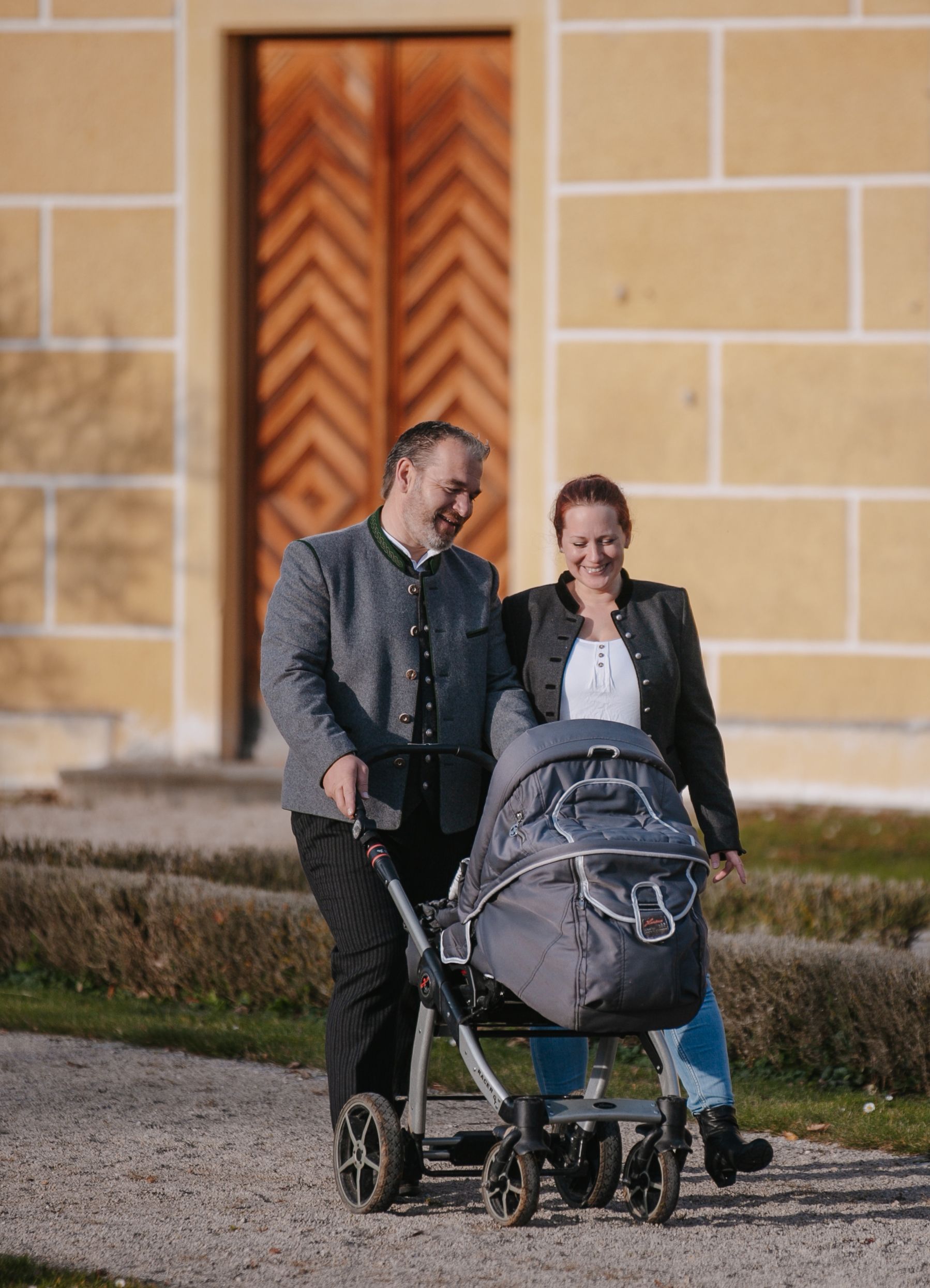 Matthias Plattner mit seiner Frau und Kinderwagen spaziert im Schlosspark. Im Hintergrund eine Holztür des Schlosses.