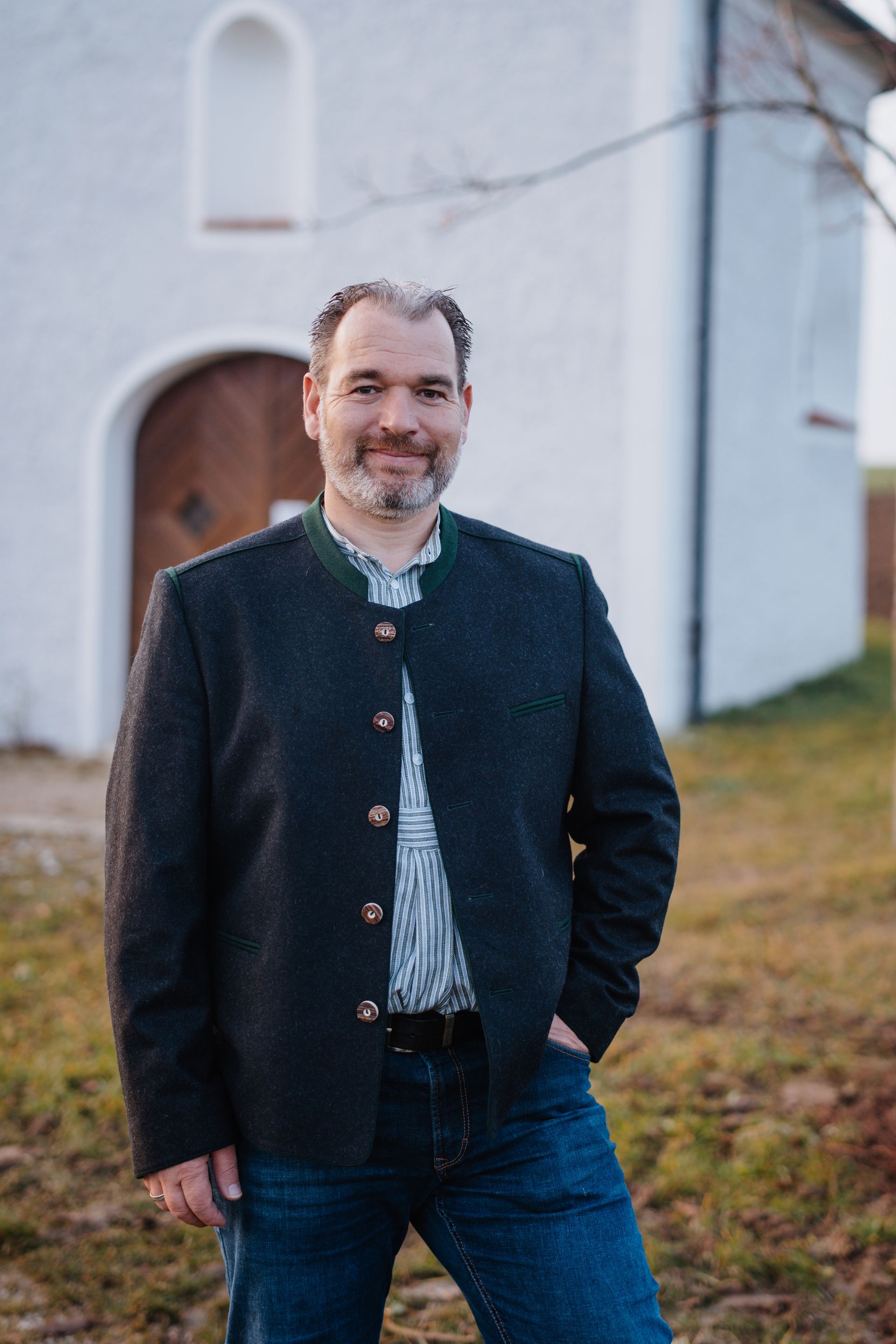Matthias Plattner in dunkler Jacke und Jeans posiert vor einem kleinen weißen Gebäude.