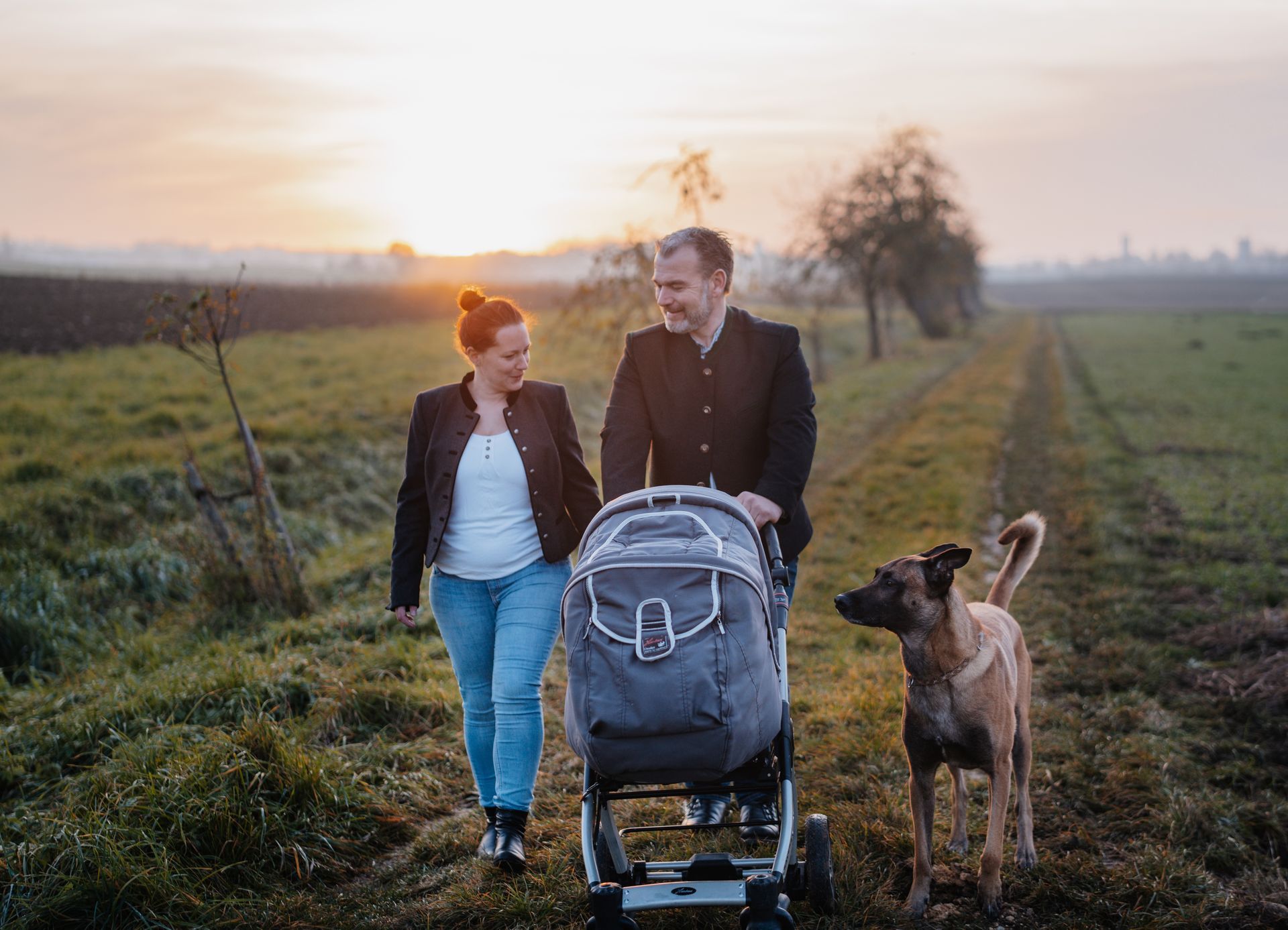 Mathias Plattner mit Frau schiebt den Kinderwagen auf einem Feldweg, ein Hund begleitet sie, im Hintergrund Sonnenuntergang.
