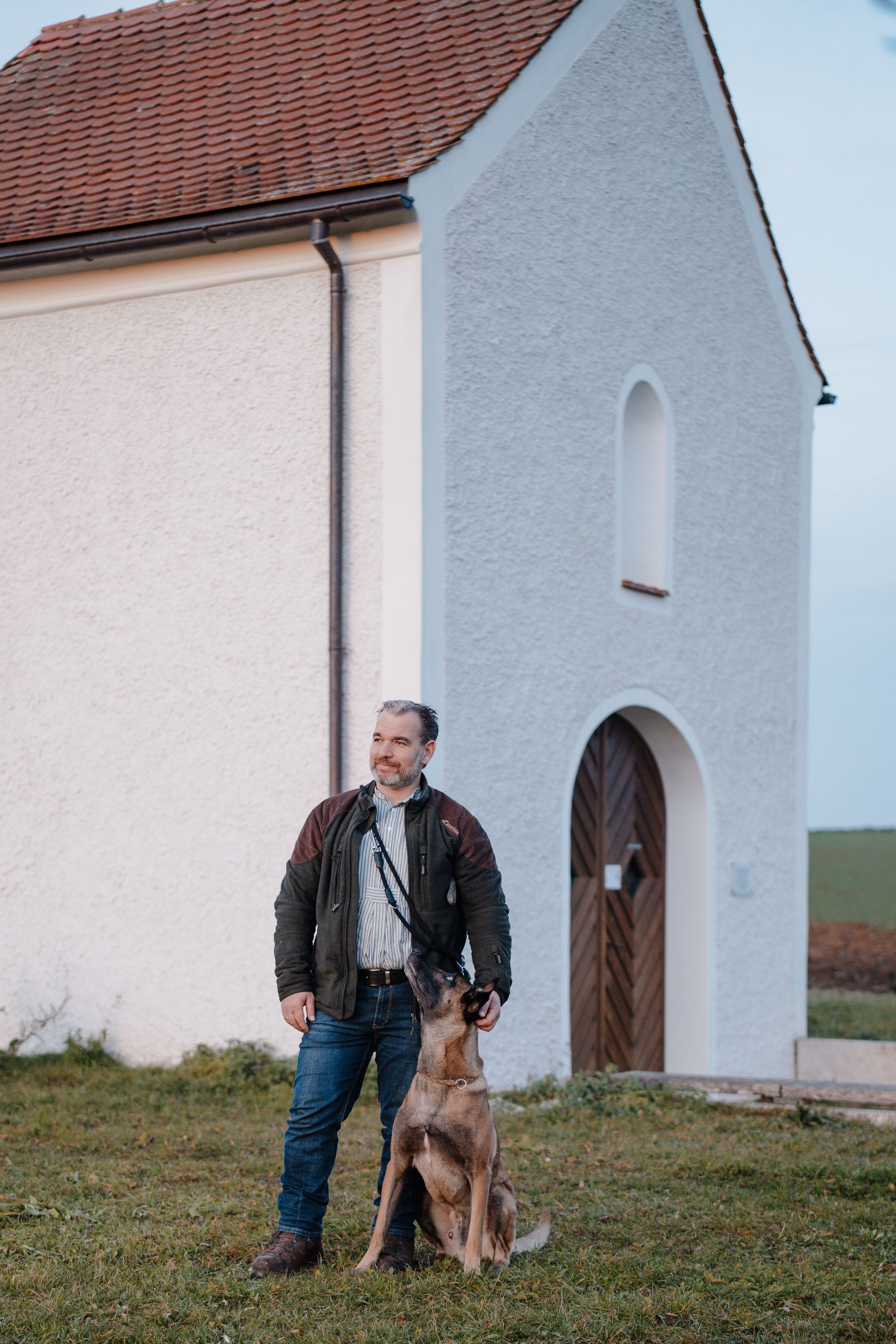 Matthias Plattner mit Hund steht im Freien vor einem weißen Gebäude mit rotem Dach.