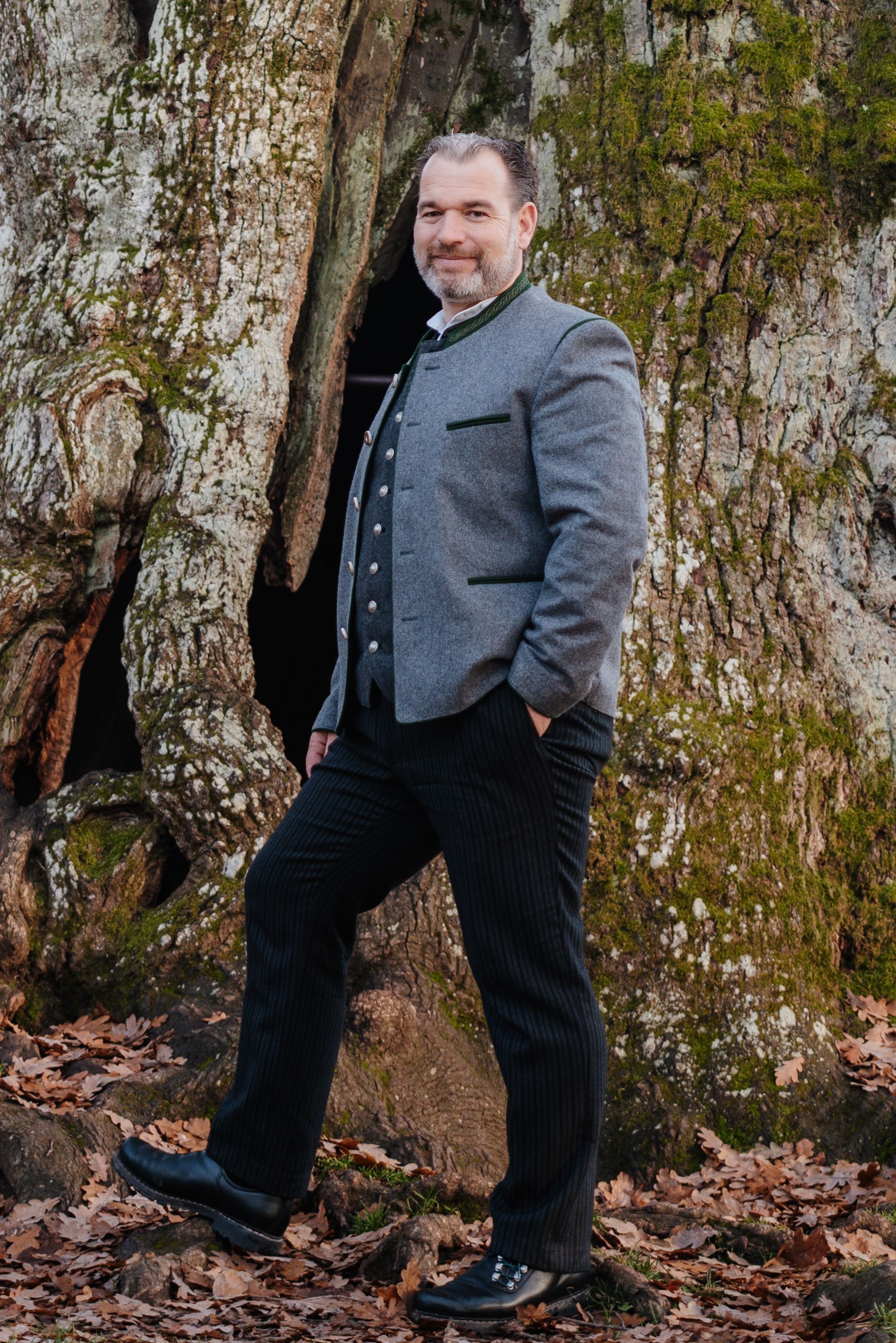 Matthias Plattner in traditioneller grauer Jacke und schwarzer Hose posiert neben einem großen Baum.