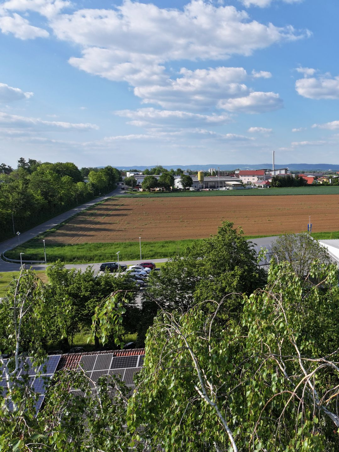 Blick von oben auf ein Feld, eine Straße zum Feuerwahrhaus und das Gewerbegebiet unter blauem Himmel mit flauschigen Wolken.
