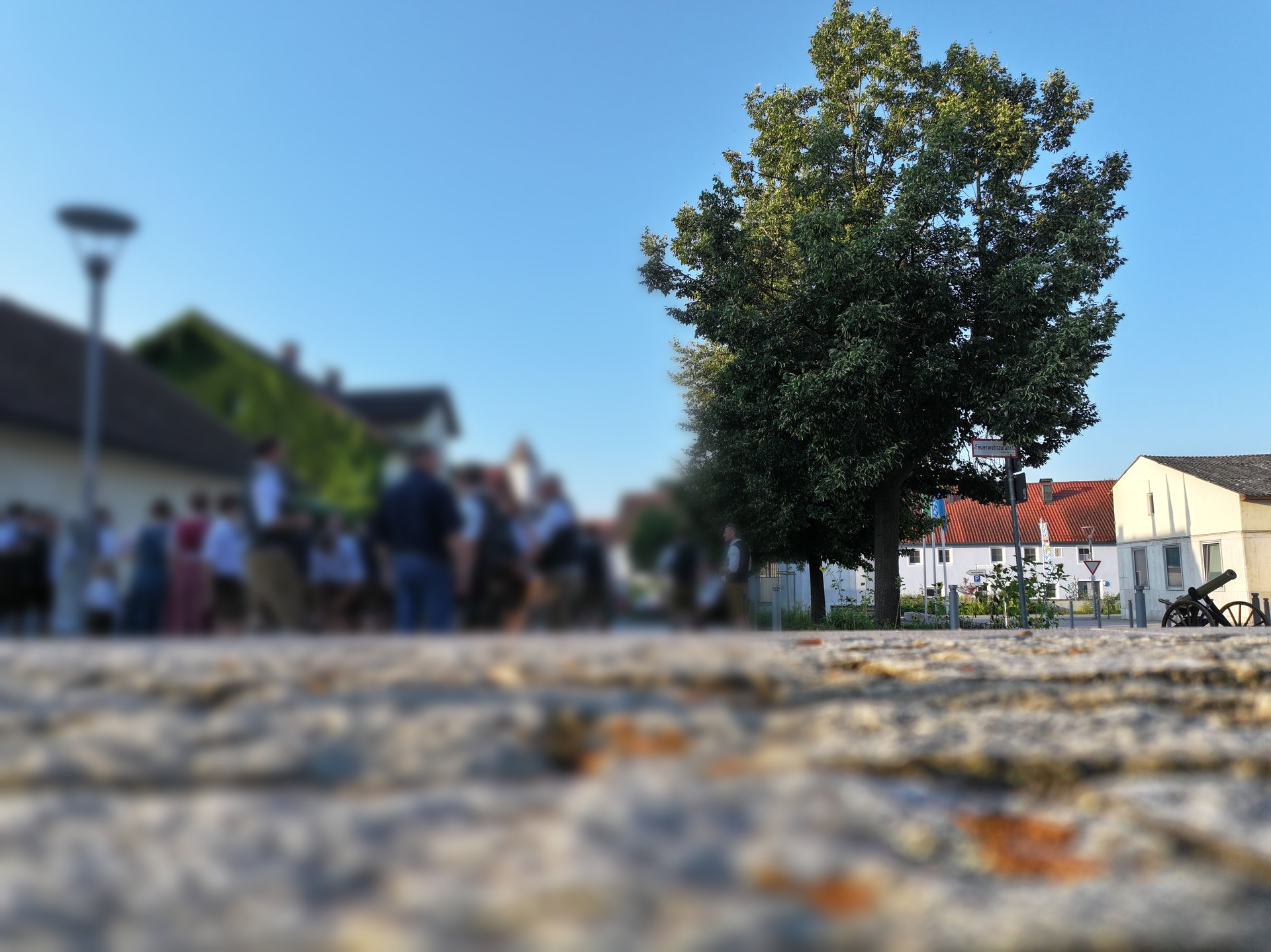 Der Dorfplatz in Alteglofsheim: Kopfsteinpflasterweg, Baum und Gebäude, im Hintergrund sind Menschen verschwommen zu erkennen