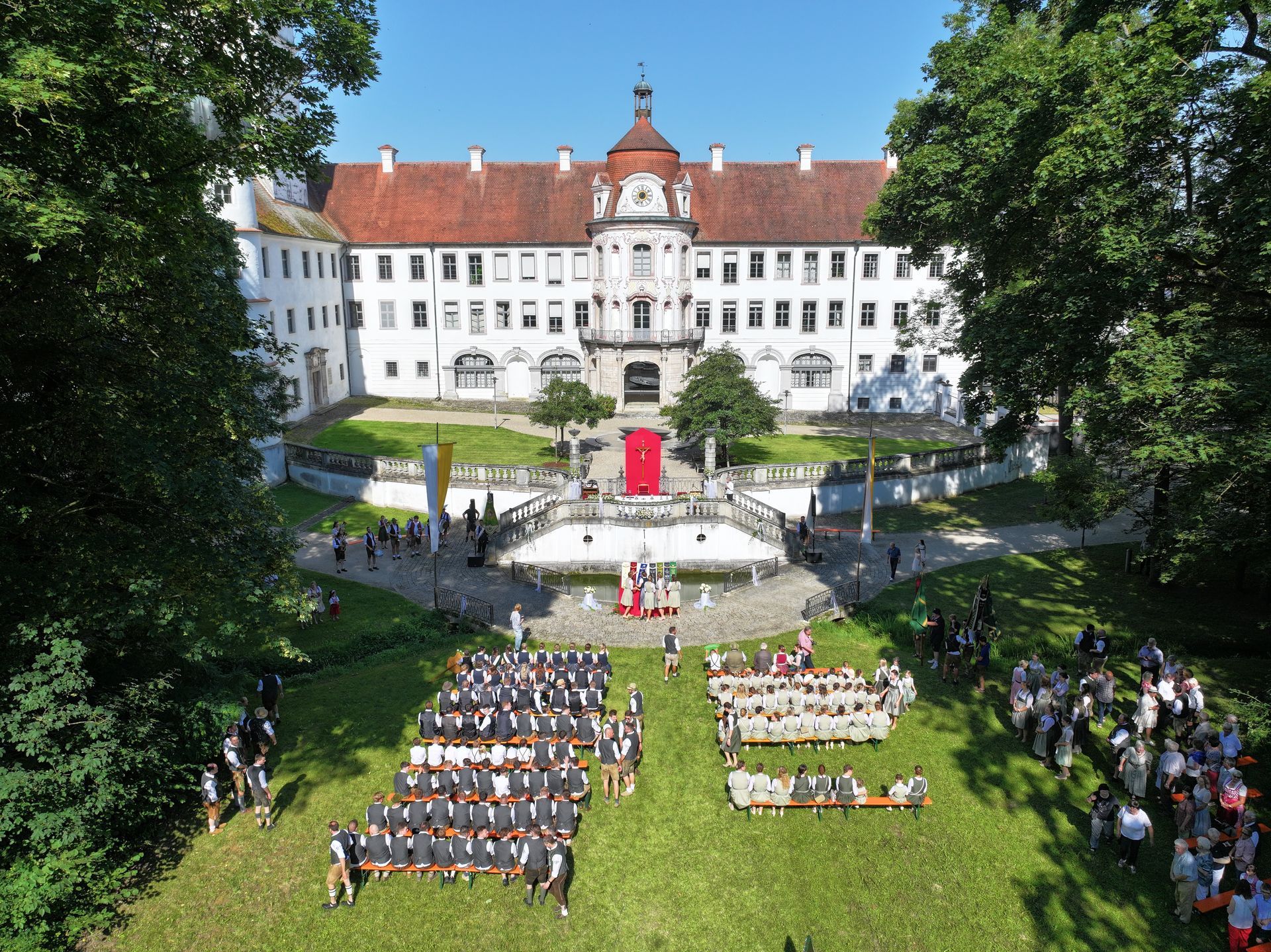 Fahnenweihe im Schlosspark des Schlosses Alteglofsheim, Zeremonie auf dem Rasen. Menschen in dunklen Gewändern, Bäume.