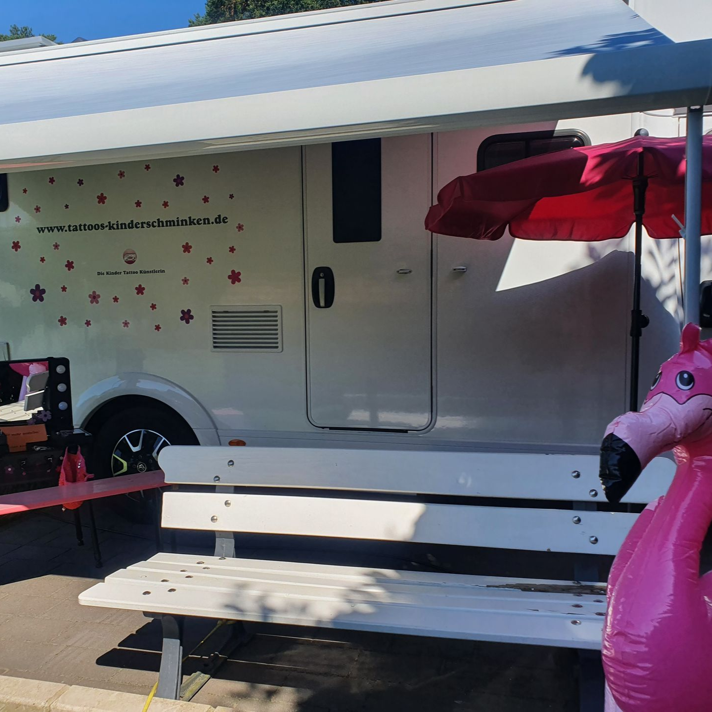 Weißes Wohnmobil mit rosa Dekorationen; Außensitzgruppe mit Bank, Sonnenschirm und Flamingo-Luftmatratze.