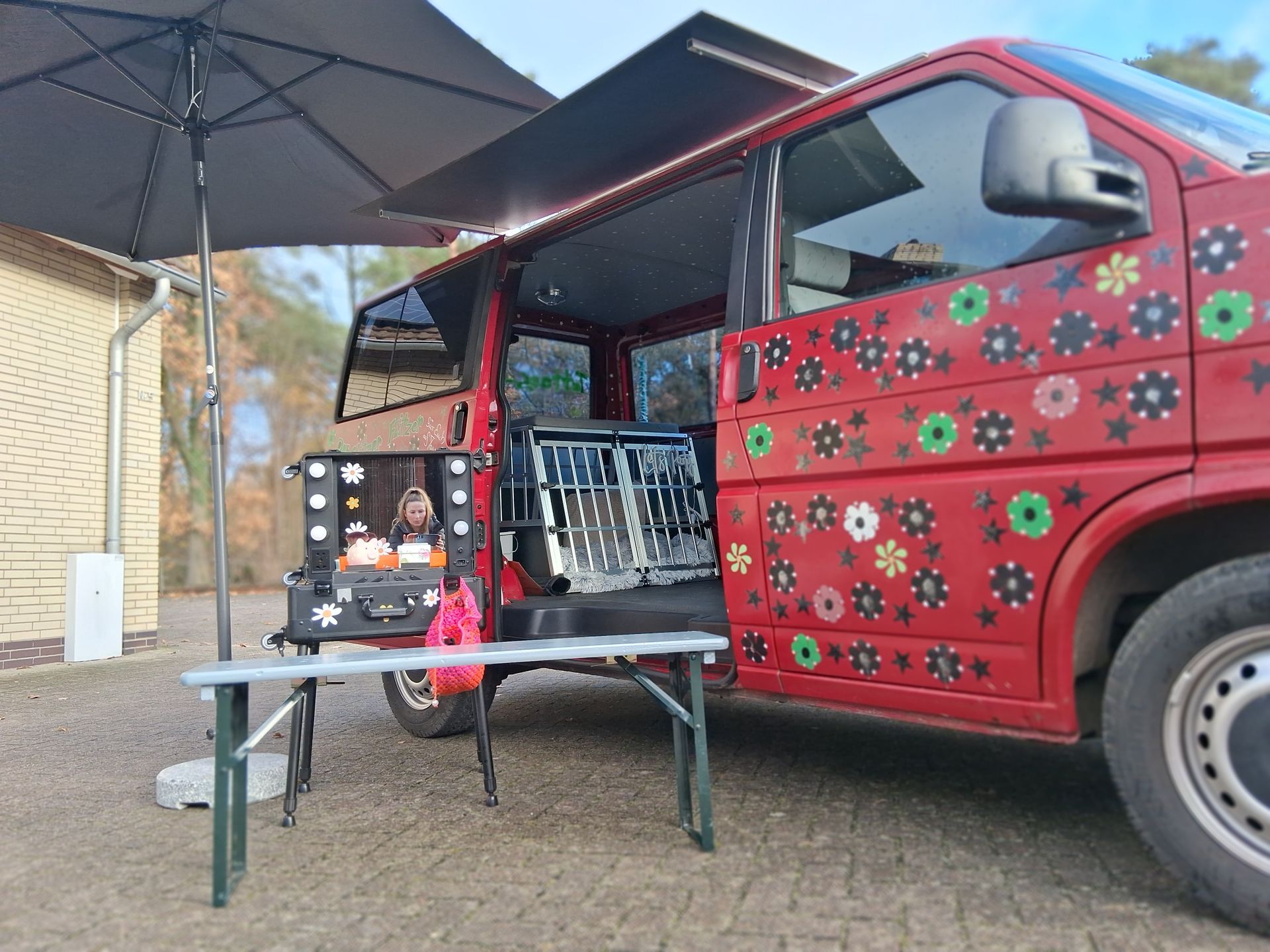 Roter Lieferwagen mit offener Seite, auf dem Waren ausgestellt sind; Tisch und Sonnenschirm sind im Freien aufgebaut.