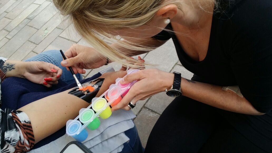 Eine Person malt ein buntes Muster auf den Arm einer anderen; sie hält eine Farbpalette in der Hand. Draußen.