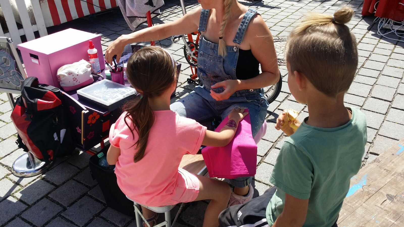 Frau in Latzhose mit zwei Kindern an einem kleinen Verkaufsstand, eines der Kinder hält einen rosa Gegenstand.