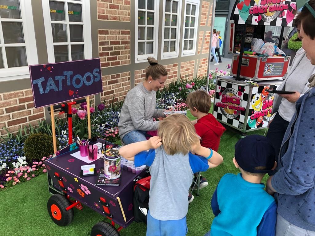 Eine Frau verpasst Kindern an einem mit Blumen und einem „Tattoos“-Schild geschmückten Stand im Freien temporäre Tattoos.