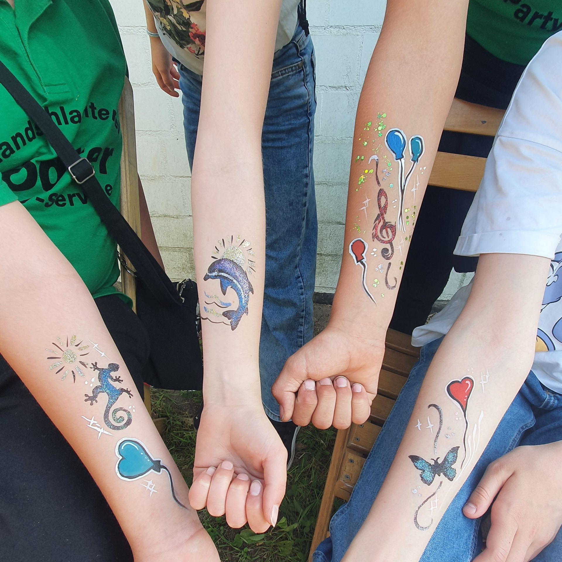 Vier Arme mit temporären Tattoos, darunter ein Delfin, Noten und Luftballons.