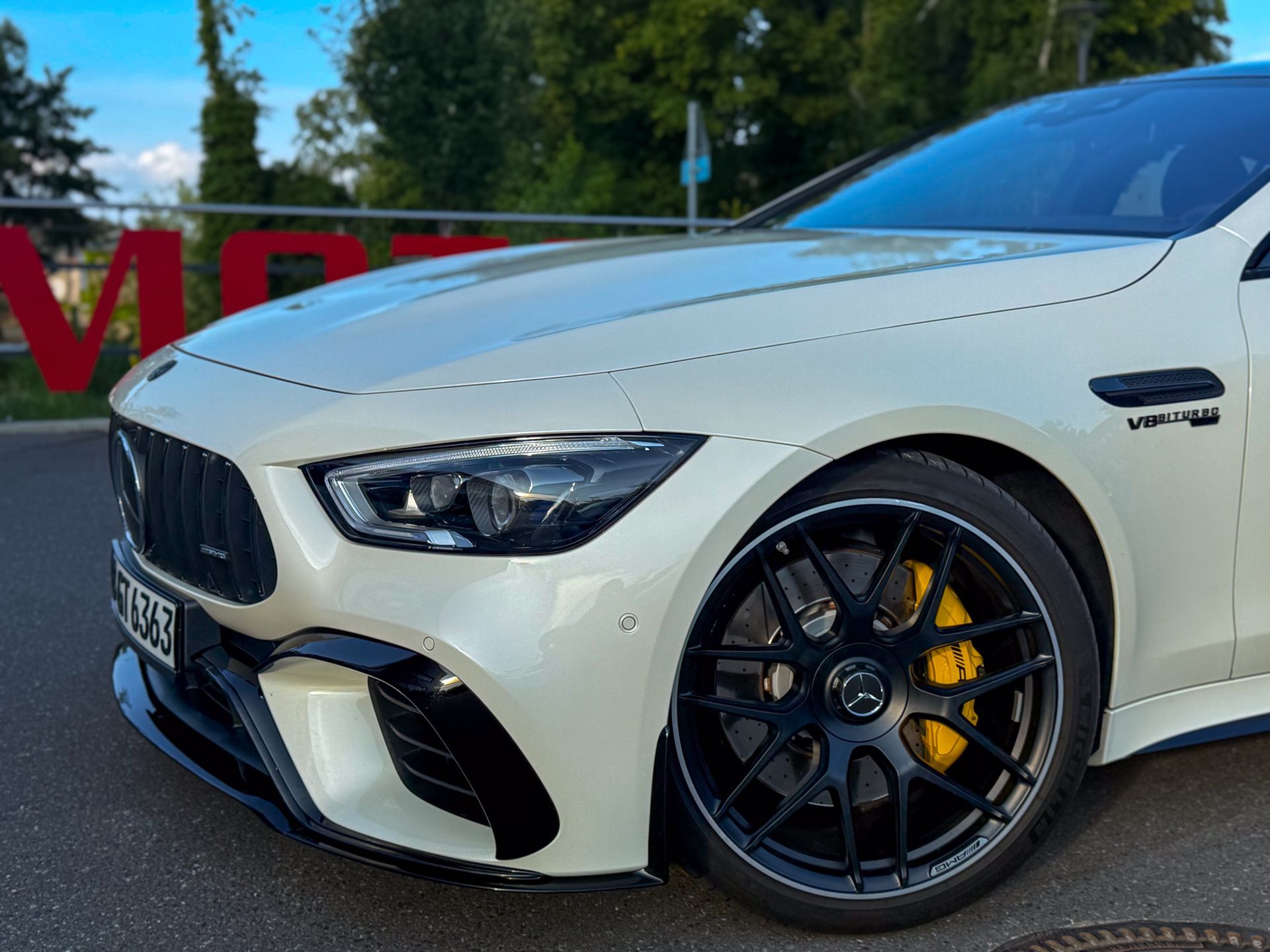 Nahaufnahme der Frontpartie eines weißen AMG GT63s von vorne links, vor einem Hintergrund aus Bäumen und Himmel jetzt mieten