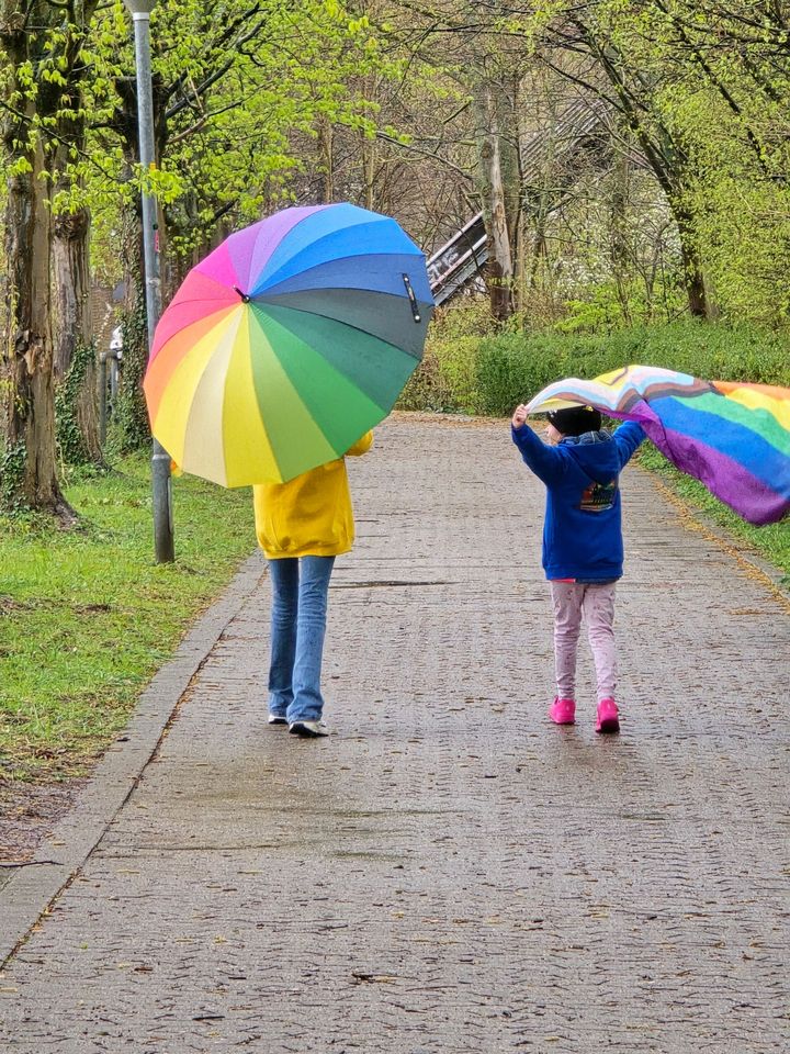 Zwei Personen spazieren mit Regenbogen-Regenschirmen auf einem Parkweg in einer farbenfrohen, grünen Umgebung.