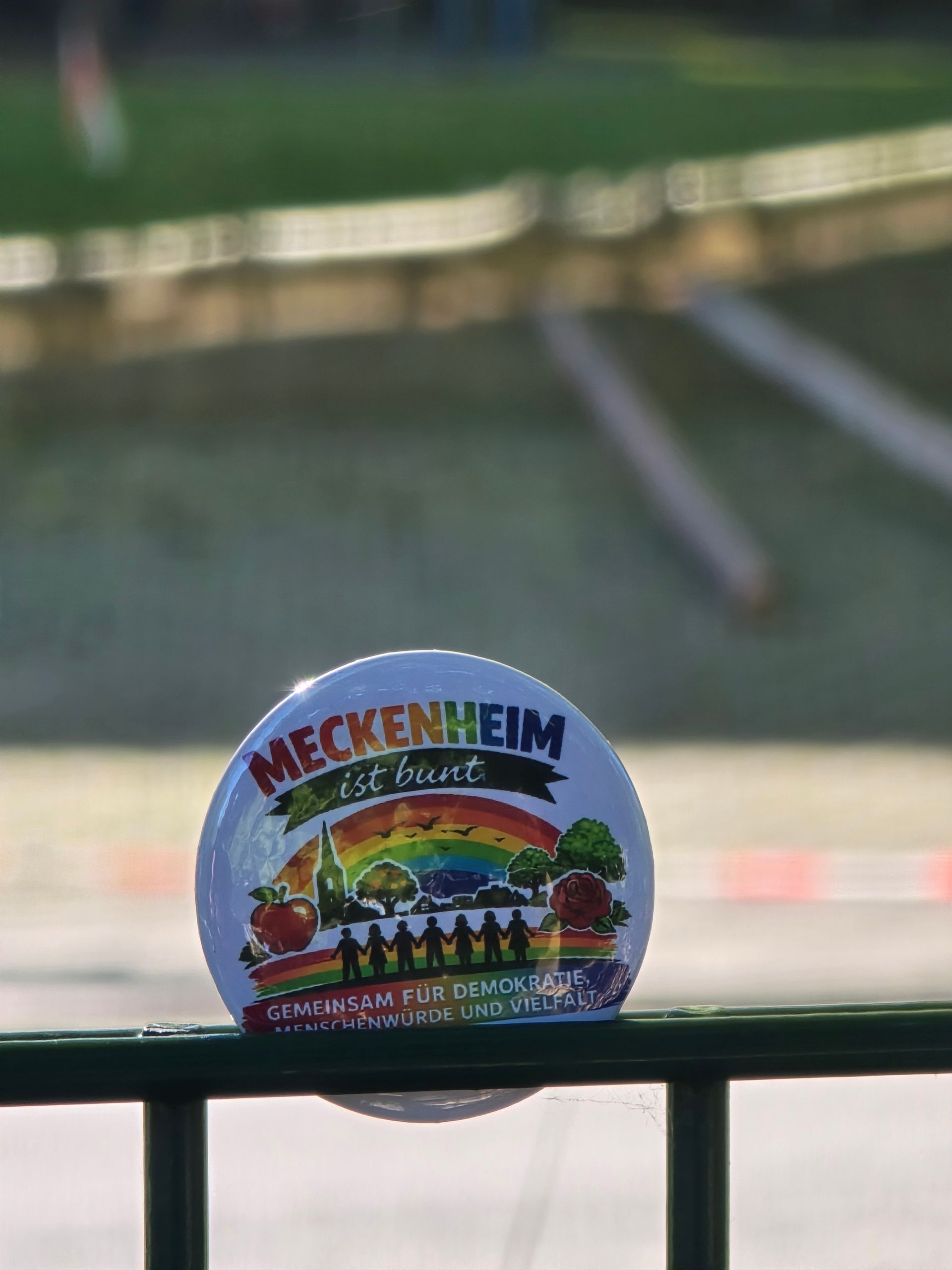 Runder Anstecker mit der Aufschrift „Meckenheim ist bunt“ an einem Geländer vor verschwommenem grünem Außenhintergrund.